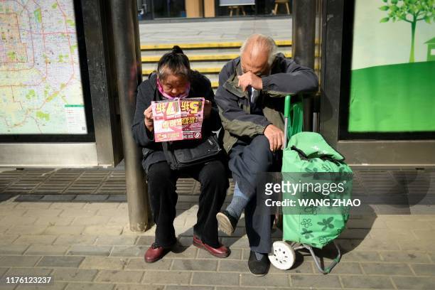 An elderly woman reads a promotional leaflet sitting next an elderly man at a bus station in Beijing on October 16, 2019. - China's economy expanded...