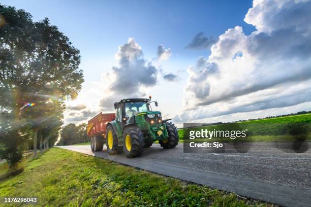 john deere traktor schleppt einen kipper anhänger auf einer landstraße zwischen landwirtschaftlichen feldern - john deere tractor stock-fotos und bilder