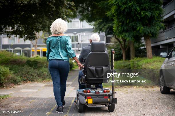 62 year old daughter walking next to 91 year old father in mobility scooter - 63 year old female - fotografias e filmes do acervo