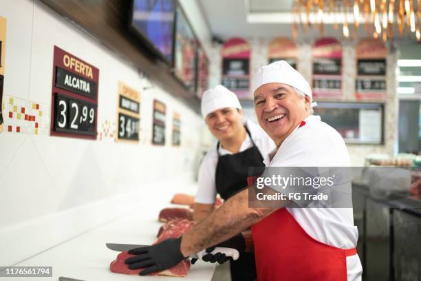 portrait of a butcher cutting a piece of red meat in butchers shop, colleague in background - apron gloves stock pictures, royalty-free photos & images