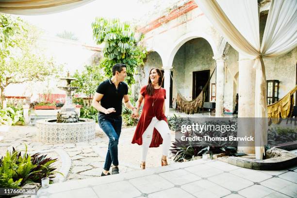 smiling couple holding hands while walking up steps in courtyard of villa - return to paradise stock pictures, royalty-free photos & images