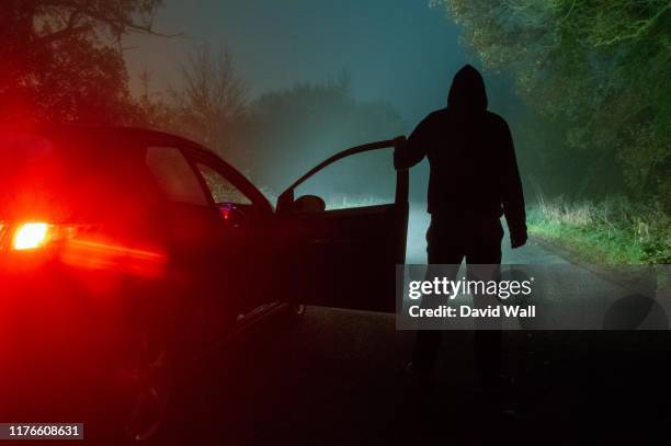 a spooky, mysterious hooded figure, standing next to a car with the door open. looking down a moody, foggy, road at night - woods at night stock-fotos und bilder