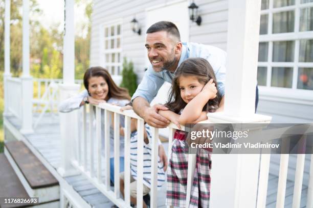 famiglia con due bambini appoggiati alla recinzione di fronte alla loro casa - family porch foto e immagini stock