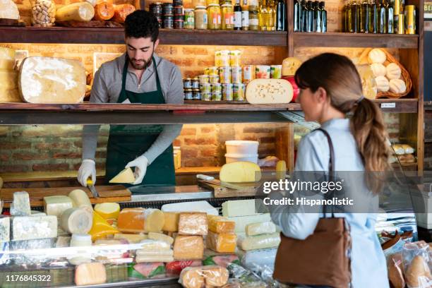 latijns-amerikaanse mannelijke verkoopmedewerker snijden van een stukje kaas voor vrouwelijke klant bij de delicatessen - french cheese stockfoto's en -beelden