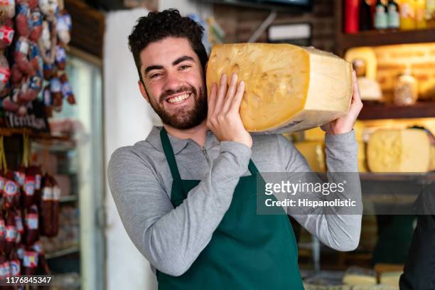 male sales clerk at a delicatessen carrying a heavy cheese on shoulder while smiling at camera very cheerfully - french cheese stock pictures, royalty-free photos & images