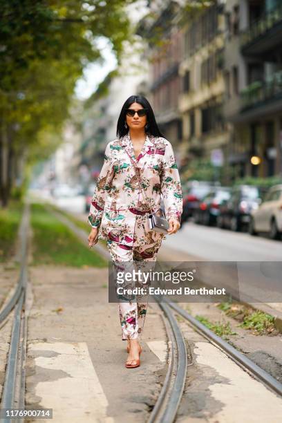 Guest wears sunglasses, earrings, beige pajamas suit with colorful cats and flowers pattern, a shiny silver bag , transparent orange vinyl sandals,...