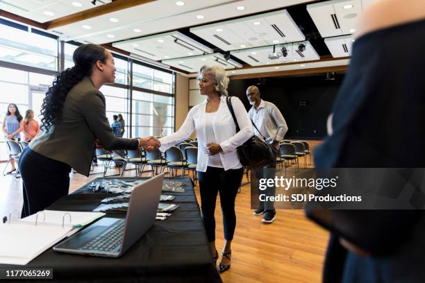 mid adult woman welcomes mature female conference attendee - diverse town hall meeting stock pictures, royalty-free photos & images