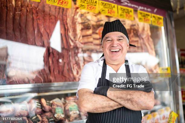 portret van een senior slager staande met armen gekruist voor een slagers winkel - butcher stockfoto's en -beelden