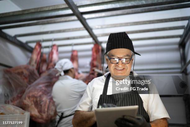 senior slager met behulp van digitale tablet om producten te analyseren in een vlees locker - koelkast stockfoto's en -beelden