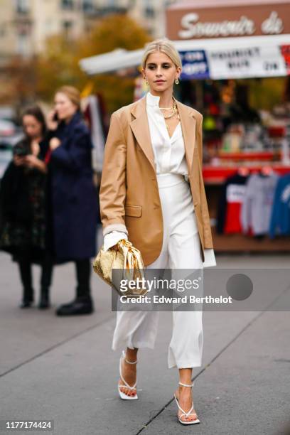 Caroline Daur wears earrings, necklaces, a camel leather oversized jacket, a white shirt, white wide-legs crop pants, white sandals, a shiny gold...