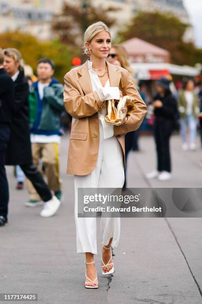 Caroline Daur wears earrings, necklaces, a camel leather oversized jacket, a white shirt, white wide-legs crop pants, white sandals, a shiny gold...