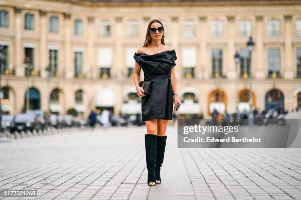 Maria Rizzo wears earrings, sunglasses, a black off-the-shoulder dress, a black Yves Saint Laurent clutch, black sued knee-high boots with shiny gold...