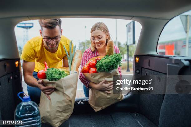 feliz pareja joven poniendo comestibles en un coche en el estacionamiento - trunk fotografías e imágenes de stock