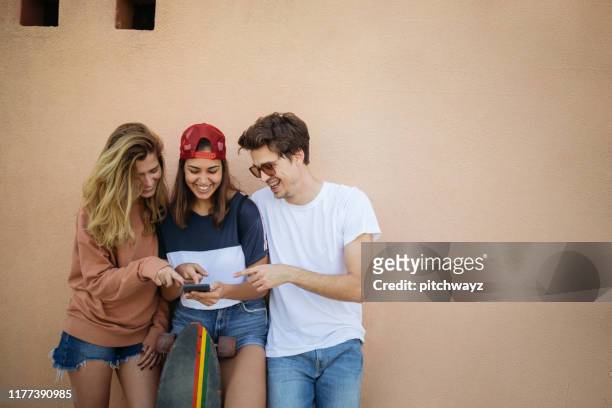 drei jugendliche friands an der wand lehnen und blick auf smartphone. - 3 teenagers mobile outdoors stock-fotos und bilder