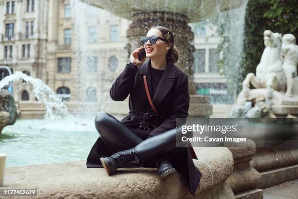 young woman sitting by the fountain and talking on the phone - leather trousers stock-fotos und bilder