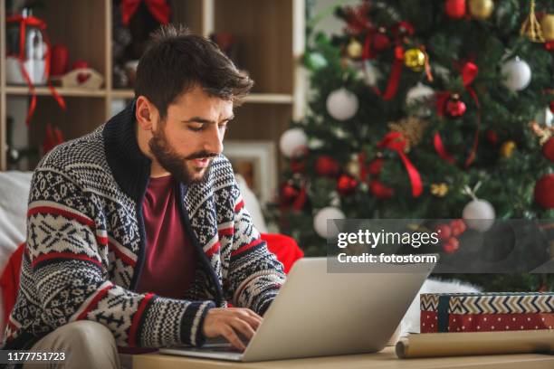 junger mann mit laptop im wohnzimmer in der nähe des weihnachtsbaumes - weihnachten laptop stock-fotos und bilder