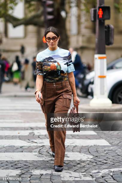 Camelia Jordana wears sunglasses, earrings, a bracelet, a Chloe top with a landscape print, brown houndstooth cargo pants, a brown crocodile Chloe...