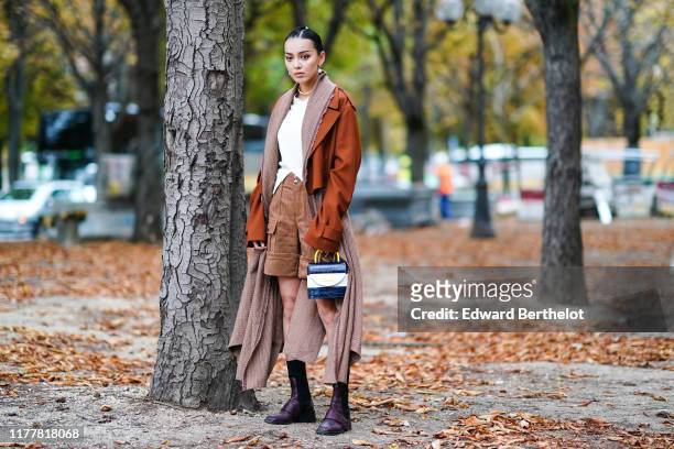 Kiwi Lee Han wears earrings, a necklace, a white V-neck top, a rust-color oversized jacket, brown houndstooth cargo shorts, a large and long...