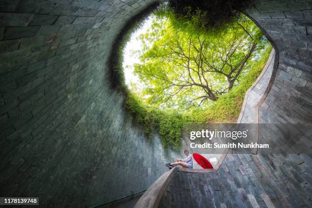 woman siting on the handrail under giant tree with red umbrella at spiral staircase of underground in tunnel crossing of road at fort canning park, singapore - fort canning stock pictures, royalty-free photos & images