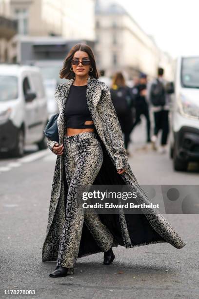 Camila Coelho wears sunglasses, a snake print pattern long coat, flare pants, a cropped top, earrings, black shoes, outside Giambattista Valli,...