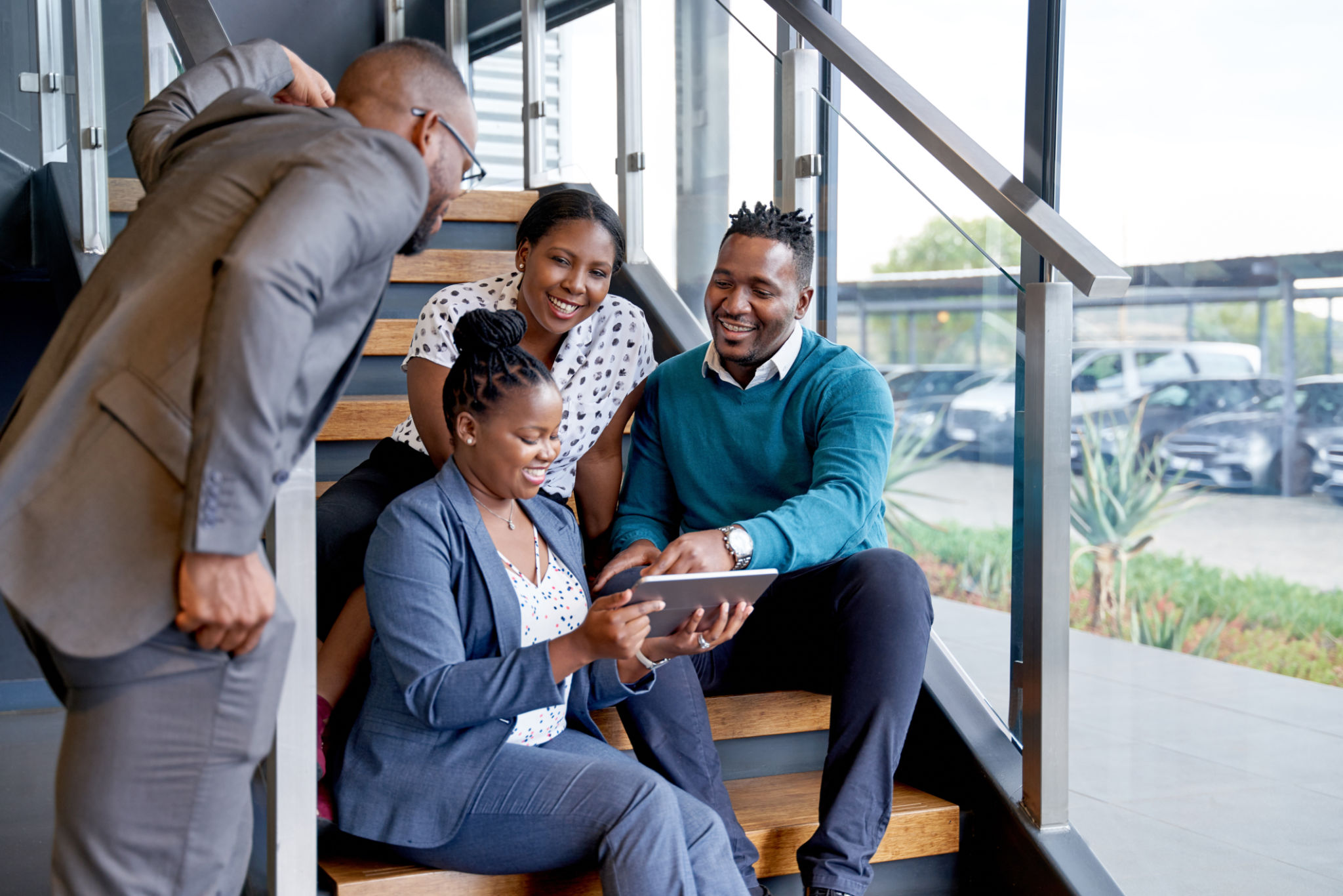 Four professional male and female coworkers sitting on staircase smiling practicing teamwork on corporate tablet Four professional male and female coworkers sitting on staircase smiling practicing teamwork on corporate tablet