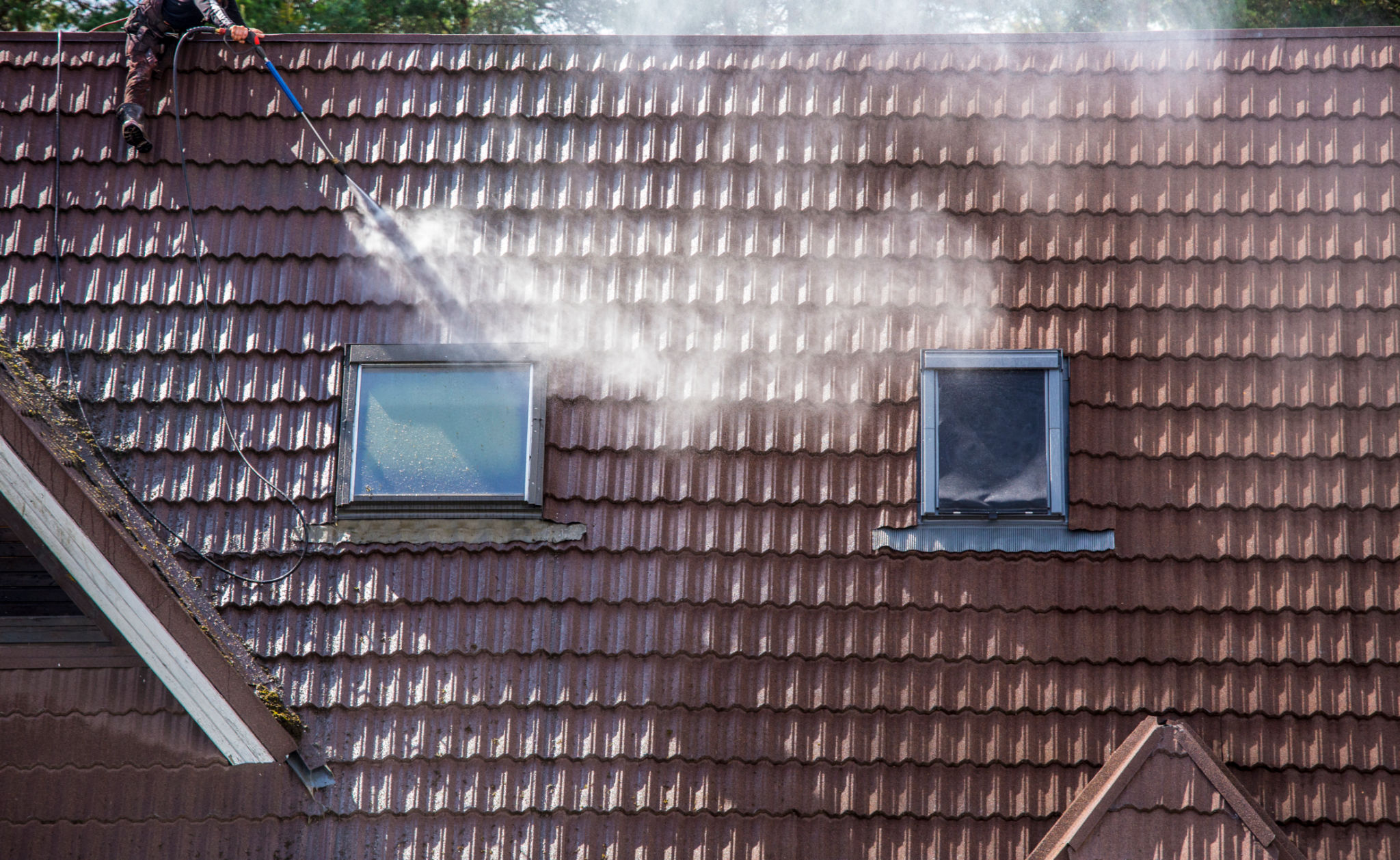 Man worker washing tilted stone roof shingles with water pressure washer to remove dirt, moss and lichen outdoors in summer. Roof cleaning service concept. Man worker washing tilted stone roof shingles with water pressure washer to remove dirt, moss and lichen outdoors in summer. Roof cleaning service concept.