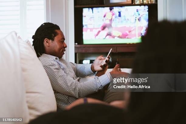 father texting whilst mother watches sport on tv - african family watching tv stockfoto's en -beelden
