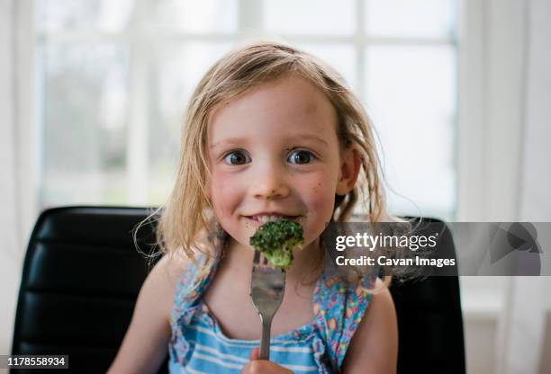 young girl eating broccoli at home with a messy face - parenting dinner kids stock-fotos und bilder