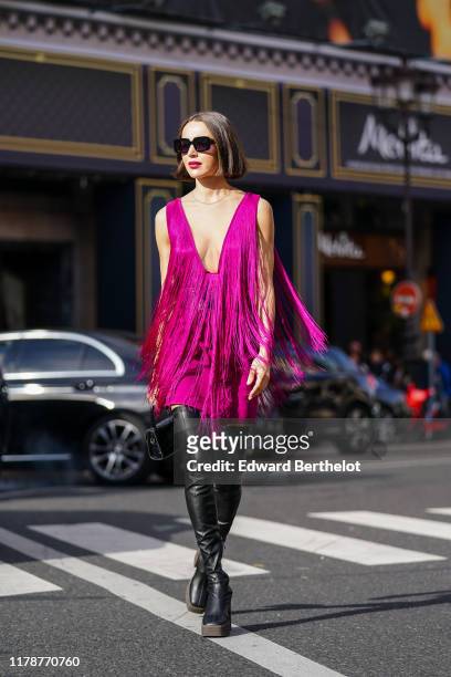 Mary Leest wears sunglasses, a fuchsia fringed mini dress with a plunging neckline, a large black belt with metallic rings, a black crocodile pattern...