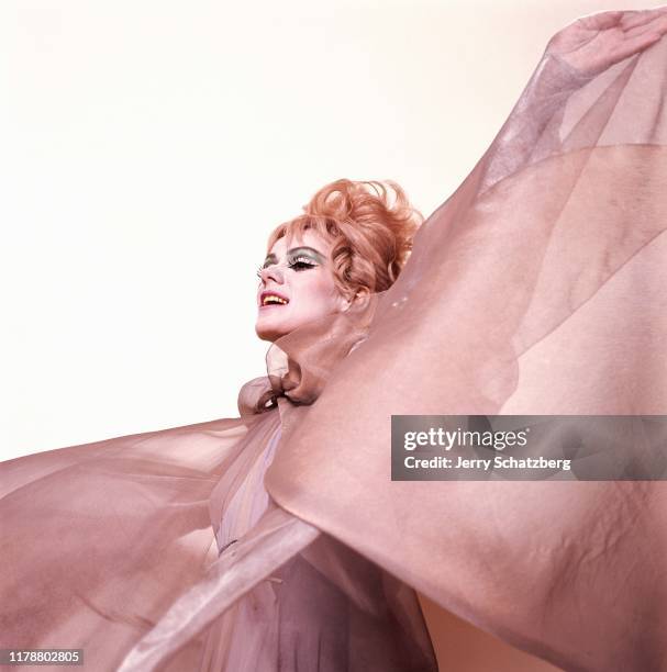 In heavy makeup and a long-sleeved, rose-colored, chiffon dress, American actress and singer Tammy Grimes raises an arm as she poses against a white...