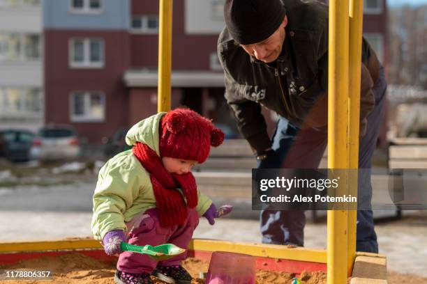 70-jähriger mann, der großvater, spielt mit seiner zweijährigen enkelin in einem sandkasten. - sandkasten stock-fotos und bilder