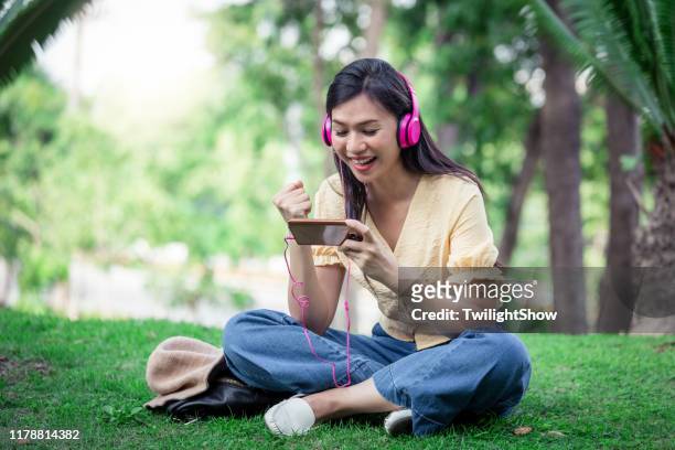 young beautiful woman sit playing the game on smartphone with pink headphones - telephone game stock pictures, royalty-free photos & images