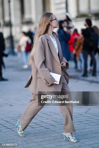 Guest wears earrings, necklaces, a white top, a light brown oversized pantsuit, blue heeled sandals, outside Louis Vuitton, during Paris Fashion Week...