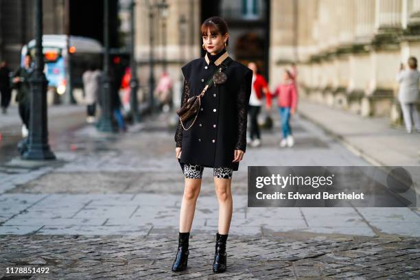 Guest wears earrings, a black lace hi-neck top, a black sleeveless mini chasuble dress with a leather collar and buttons, white and black spotted...