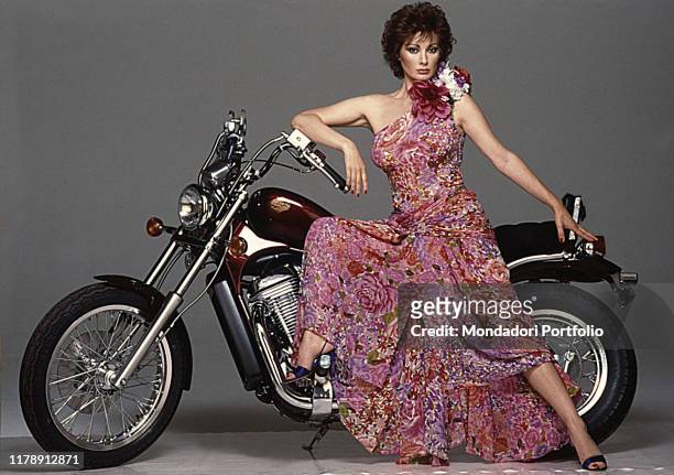 French-born Italian actress Edwige Fenech leaning against a motorcycle. 1986