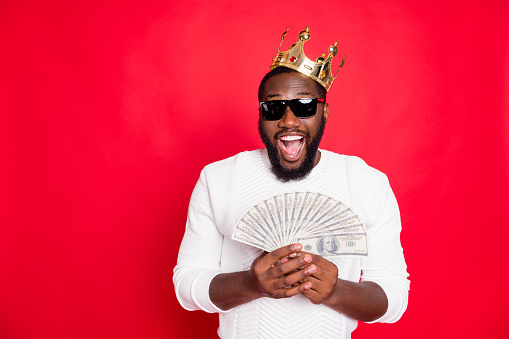Portrait of amazed surprised crazy funky dark skin man with brown hair beard hold fan money win jackpot in x-mas casino got profit become millionaire wear white jumper isolated red color background