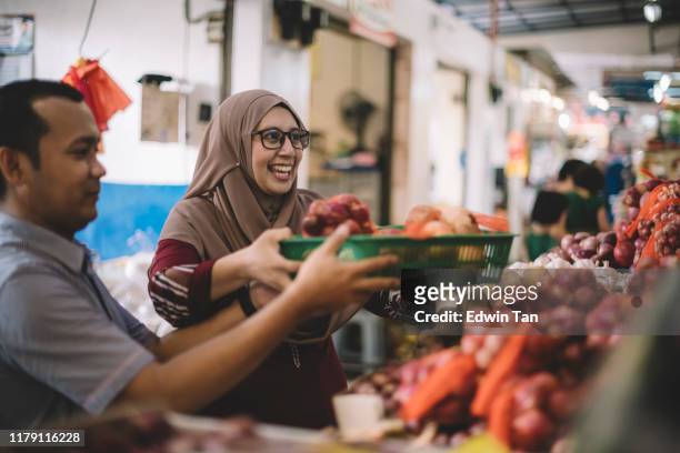 an asian malay couple buying onion . potatoes from a spice store in the market hand over the basket for price calculation