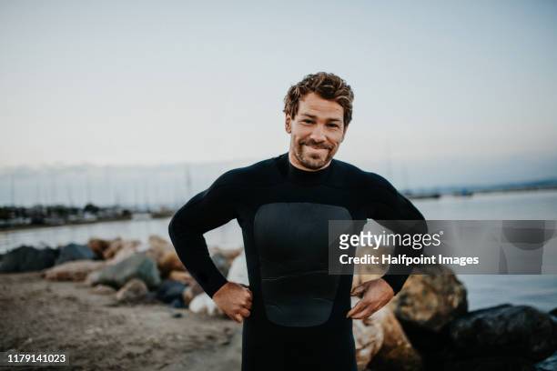 a fit mature sportsman with wetsuit standing outdoors on beach. - neoprene fotografías e imágenes de stock