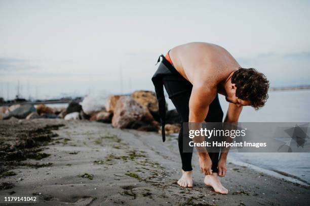 a fit mature sportsman putting on wetsuit outdoors on beach. - neopren stock-fotos und bilder