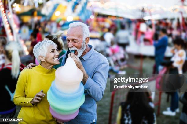 happy senior couple eating cotton candy at amusement park. - traveling carnival stock pictures, royalty-free photos & images