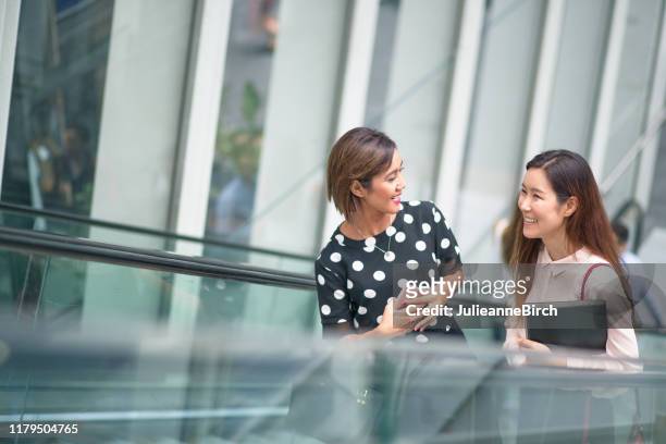 zwei asiatische frauen sprechen auf rolltreppe auf dem weg zur arbeit in der stadt - rolltreppe von oben stock-fotos und bilder