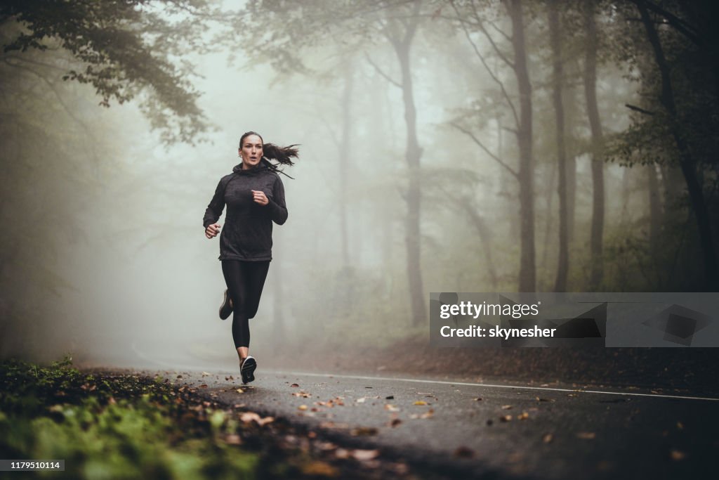 霧の森の中で道路をジョギングしている若い運動女性。