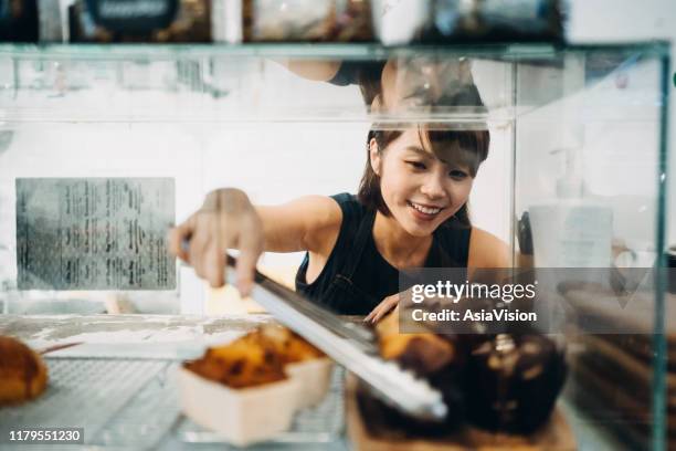 cheerful young asian woman working at a bakery and serving fresh bakery from the display cabinet - coffee shop owner stock pictures, royalty-free photos & images