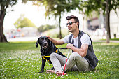 Young blind man with white cane and guide dog sitting in park in city.