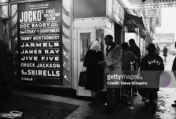View of people line up to purchase tickets at the Apollo Theater box office window, New York, New York, 1961. A poster advertises performances by the...