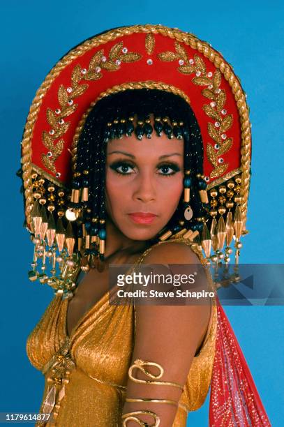 Portrait of American actress Diahann Carroll as she poses, in an elaborate headdress and a gold lame dress, against a blue background, Los Angeles,...