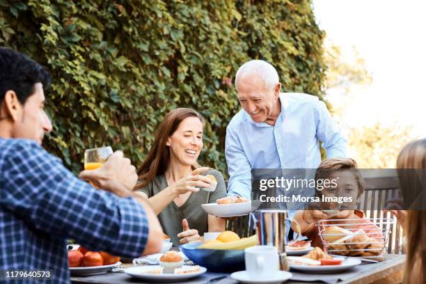 senior man passing muffins to family in backyard - standing table outside stock pictures, royalty-free photos & images