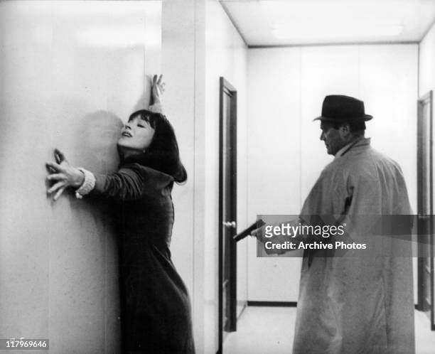 Eddie Constantine pointing a gun towards Anna Karina stretching her arms against the wall in a scene from the film 'Alphaville', 1965.