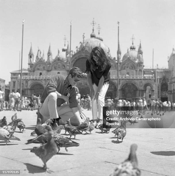 French actor Jean-Paul Belmondo, wearing a striped shirt, a sweater and sunglasses, crouched while feeding pigeons with his wife Elodie standing next...