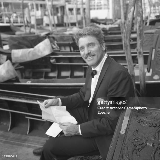 American actor Burt Lancaster, wearing a tuxedo and a bow tie and reading a letter, sitting on a gondola stationary among other gondolas, Venice 1962.